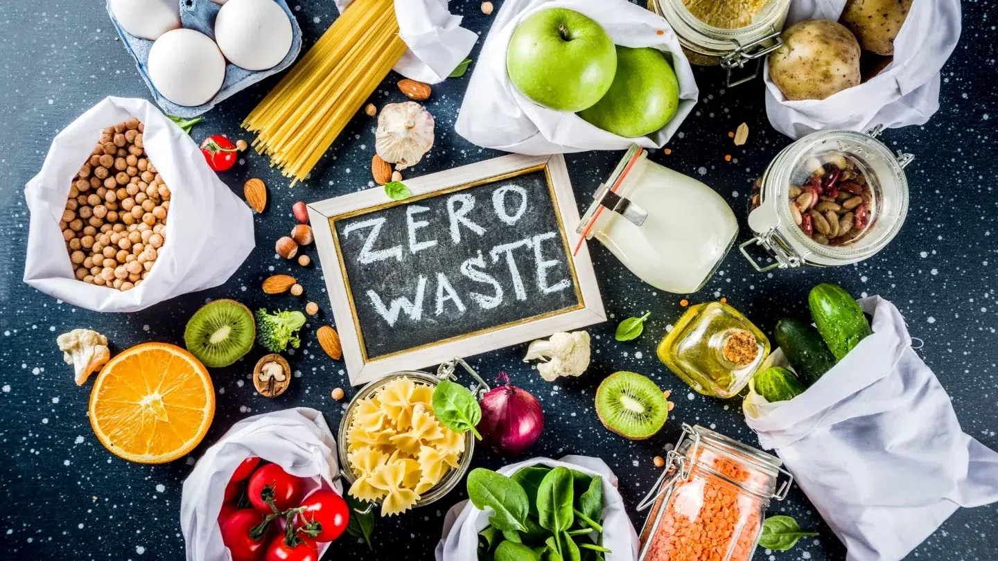Pizarra con la inscripción Zero Waste rodeada de alimentos frescos, tarrros de vidrio y bolsas reutilizables, representando el concepto y filosofía del estilo de vida residuo cero