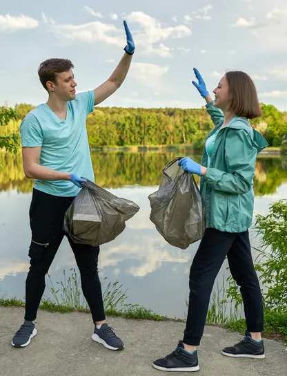 Hombre y mujer con guantes chocando los cinco sosteniendo bolsas de basura llenas junto a un lago, celebrando el final de una sesión de plogging en pareja