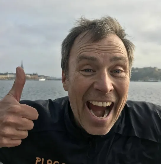 Erik Ahlström sonriente haciendo el gesto de pulgar arriba frente al agua, vistiendo una sudadera con la palabra plogging, transmitiendo la energía positiva del movimiento