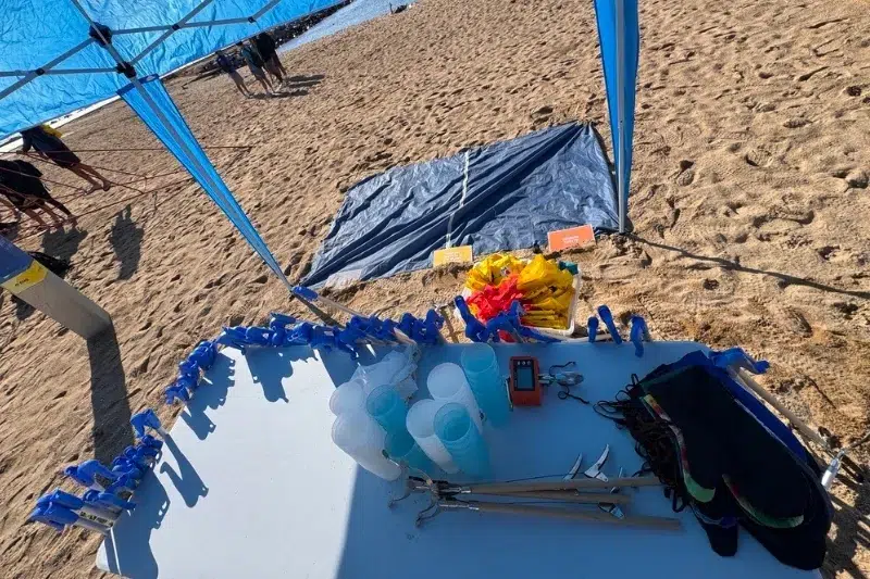 Mesa con pinzas, guantes, vasos y materiales preparados para una limpieza de playa organizada por Go Zero Waste, con carpas azules y arena de fondo
