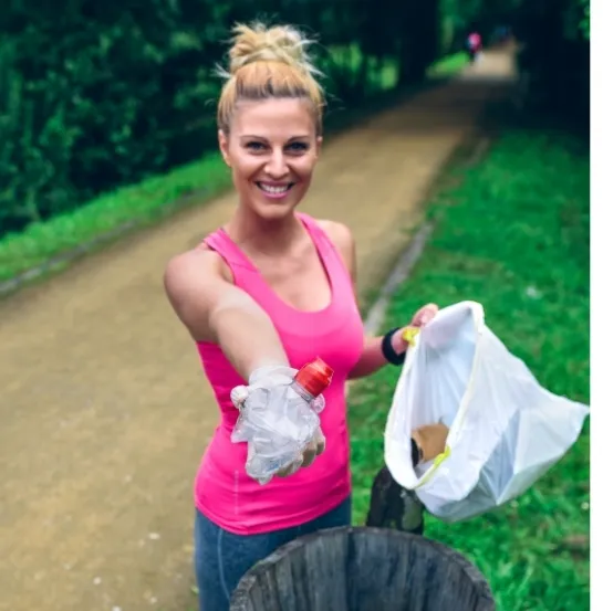 Mujer sonriente con ropa deportiva mostrando una botella de plástico recogida y una bolsa de basura junto a una papelera en un parque, practicando plogging