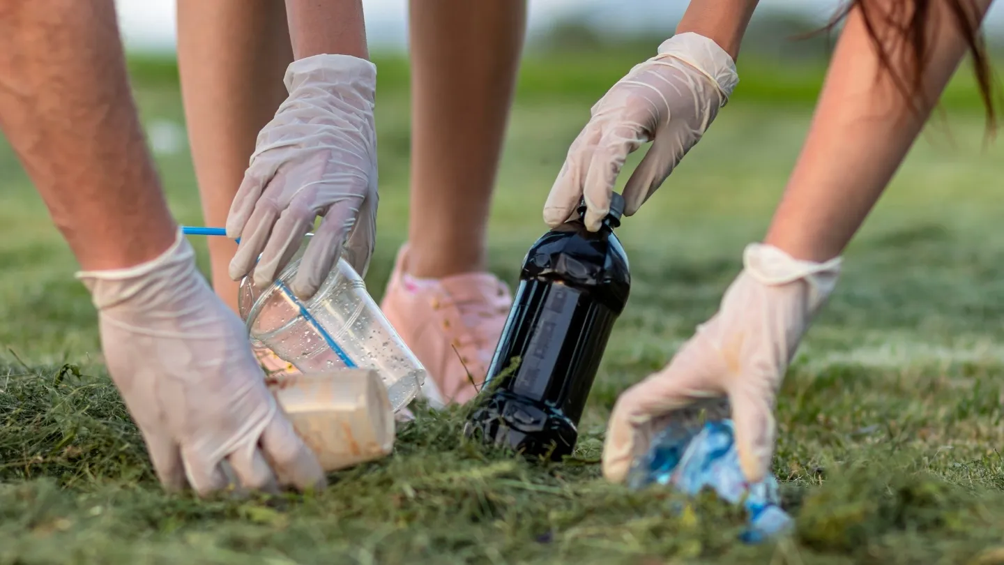 Manos con guantes recogiendo vasos de plástico, una lata y residuos en el césped, ilustrando la práctica del plogging combinando deporte y limpieza ambiental