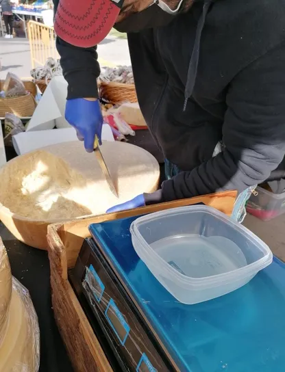 Comerciante cortando una pieza de queso para servirla en un envase reutilizable traído por el cliente
